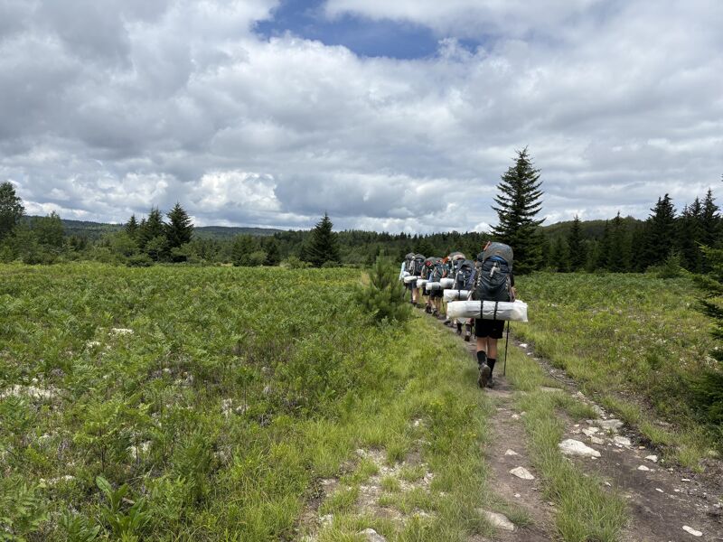 A group of people are hiking on a dirt path through a grassy field. They are wearing backpacks and carrying sleeping bags. The sky is cloudy and there are trees in the background. The hikers appear to be trekking through a natural environment, possibly on a backpacking trip or outdoor adventure.